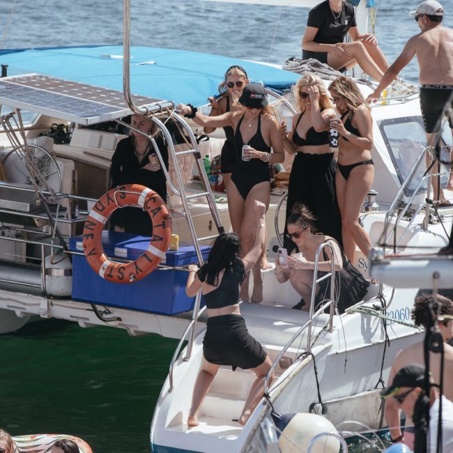 A group of people in swimwear are on a catamaran party in Sydney, with some standing on the deck holding drinks. Others sit or move around, enjoying the backdrop of water and other boats.