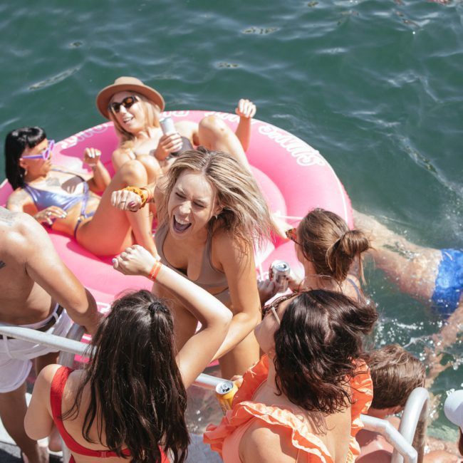 A group of people enjoys a sunny day by the water on a boat. Some are lounging on a large pink float, while others swim and socialize near the boat's edge, embracing the fun of a private yacht charter in Sydney Harbour.