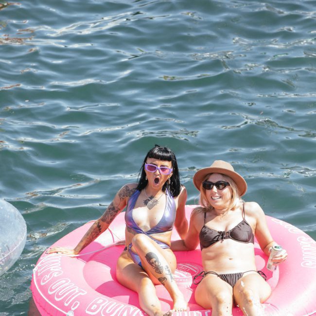 Two women in swimsuits float on a pink inflatable in water, soaking up the sun. One wears sunglasses and a hat, while the other sports tattoos and sunglasses. Nearby, the view of luxury yacht hire Sydney completes this perfect scene of relaxation.