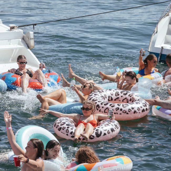 A group of people relax and enjoy themselves on inflatable pool floats in the water between two boats on a sunny day, during a corporate boat event in Sydney Harbour.