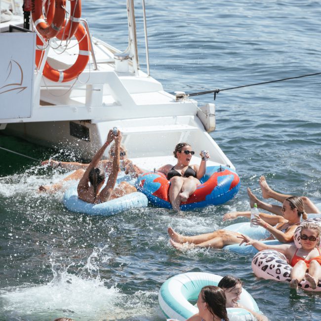 Several people are enjoying a boat party, floating on inflatable rings in the water with drinks in hand. A luxury yacht hire Sydney is docked nearby, adorned with signage featuring "Cruises.