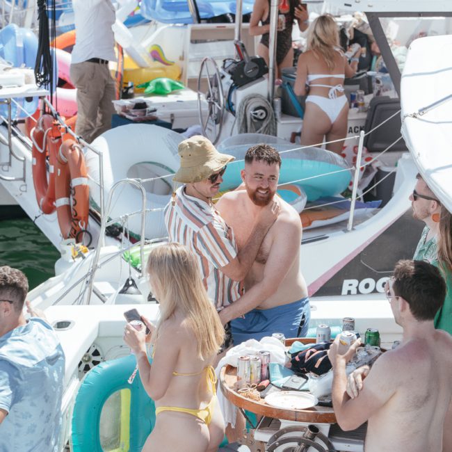 People socialize on a crowded boat deck, with two men embracing, a woman in a yellow bikini leaning on the railing, and others conversing and enjoying drinks. Various inflatable items are visible. Ideal for those looking into Sydney boat party hire.