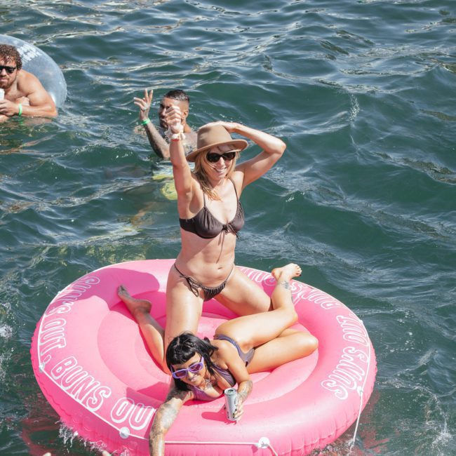 A woman in a bikini poses on a pink inflatable ring in the water while another woman lounges nearby. Two people swim close to a luxurious private yacht charter in Sydney Harbour.