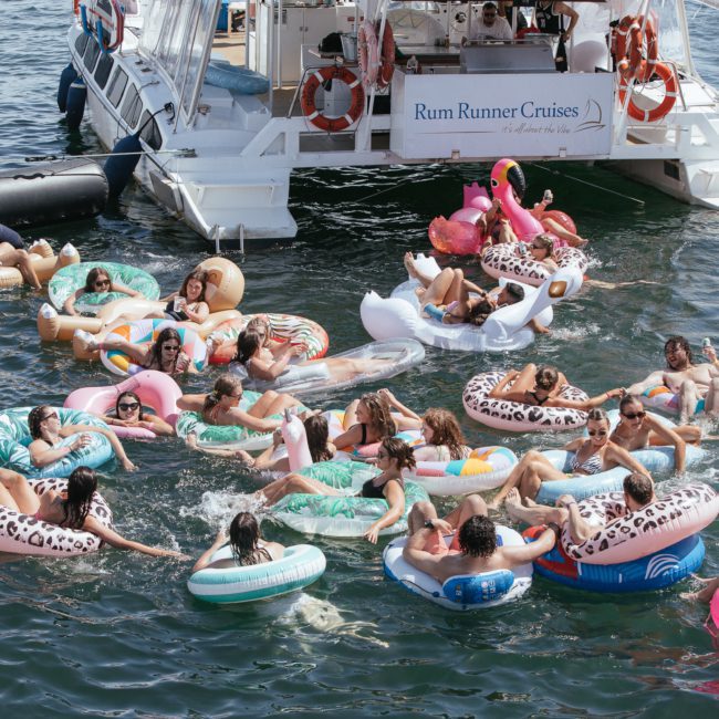 A group of people relax on various inflatable floats in the water near a boat with a "Rum Runner Cruises" sign on the back, enjoying their time with DJ boat hire Sydney.