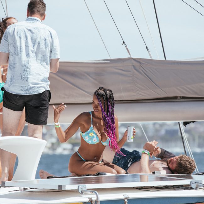A group of people in swimwear socializing on a yacht during a Sydney boat party hire. One person is sitting and holding a drink, while others stand nearby. The scene is cheerful with a backdrop of water and distant land, perfect for a private yacht charter in Sydney Harbour.