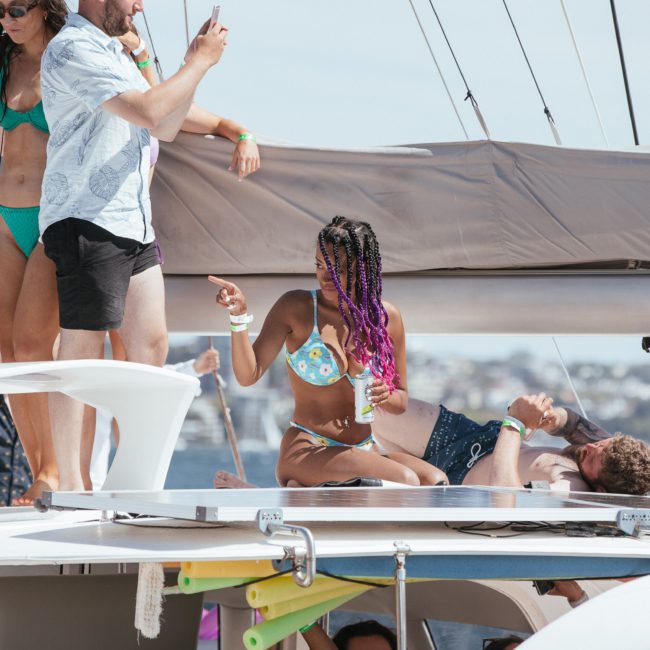 A group of people in swimwear relax on a private yacht charter in Sydney Harbour. Some are standing and socializing, while others are seated or reclining on the deck, enjoying the sunny weather.