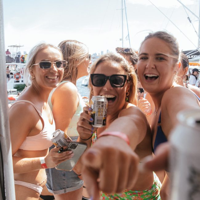 Three women in swimwear, smiling and holding drinks, on a luxury yacht hire Sydney with others in the background on a sunny day.