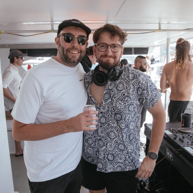 Two men on a boat, one holding a drink and the other wearing headphones near a DJ setup, enjoying a Sydney boat party hire. People and boats are visible in the background.