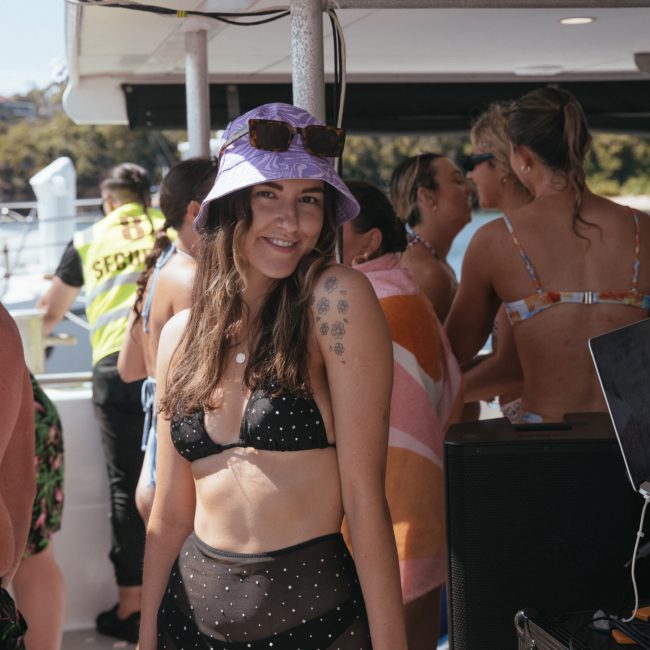 A woman in a polka-dot bikini and purple hat smiles at the camera on a boat. Other people in swimwear and a security person are visible in the background, enjoying the Sydney boat party hire. Sunlight shines brightly.