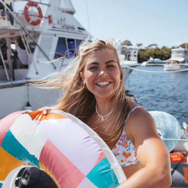 Young woman smiling while holding a colorful inflatable ring on a boat, with other boats and people visible in the background, enjoying a vibrant day made possible by DJ boat hire Sydney.