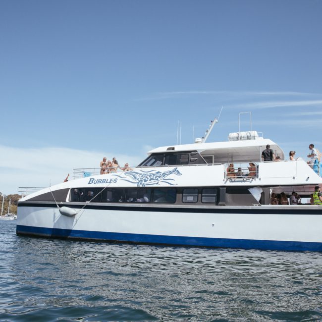 A white and blue boat named "Bubbles" with people on board floats on a calm body of water under a clear blue sky, perfect for a private yacht charter in Sydney Harbour.