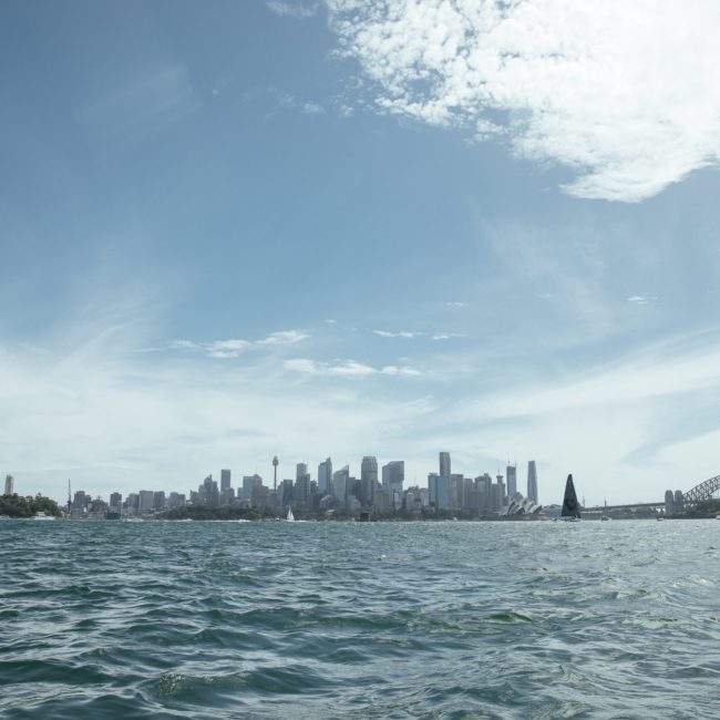 A view of the Sydney skyline with tall buildings under a partly cloudy sky, the Sydney Harbour Bridge to the right, and sailboats on the water in the foreground. It’s an ideal setting for private yacht charters on Sydney Harbour, perfect for enjoying luxury experiences amidst iconic scenes.