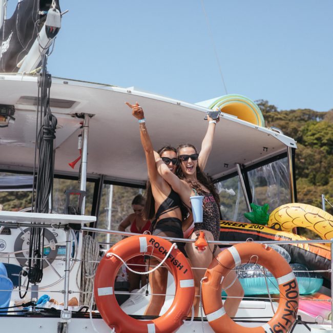 Two women in swimsuits and sunglasses cheerfully pose and wave on a private yacht charter in Sydney Harbour, surrounded by vibrant life rings and inflatable pool floats. Trees can be seen in the background.