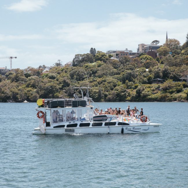 A boat filled with people is cruising on a body of water with a wooded shore and buildings in the background. A small boat is visible in the distance, perfect for corporate boat events Sydney.