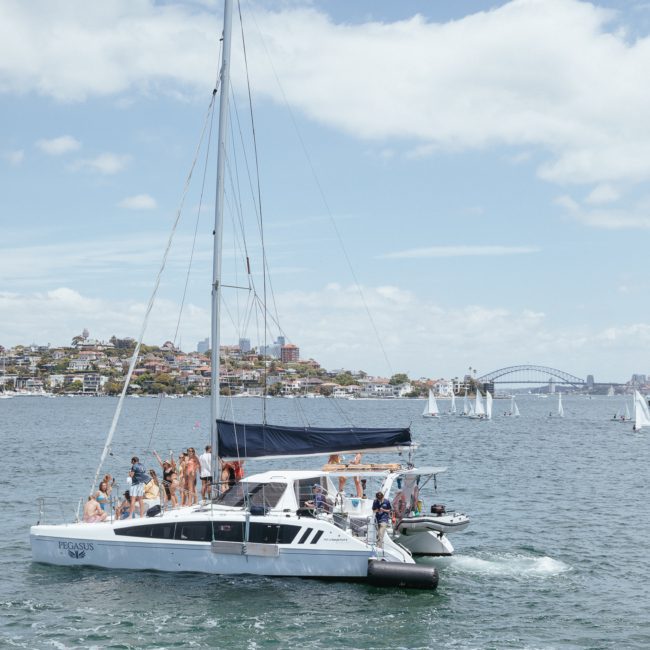 A catamaran with several people onboard sails on a sunny day. Other boats and a cityscape, including a bridge, can be seen in the background, making it perfect for a Sydney boat party hire.
