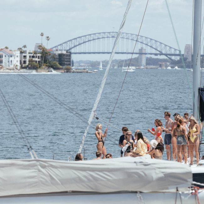 People stand on a catamaran docked in a harbor, enjoying a Sydney boat party hire with a bridge and residential buildings in the background.