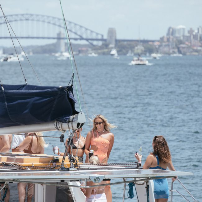 A group of people stand on a luxury yacht with a view of a large bridge and city skyline in the background. Some are holding drinks, and several boats are visible on the water, embodying the essence of a private yacht charter Sydney Harbour event.