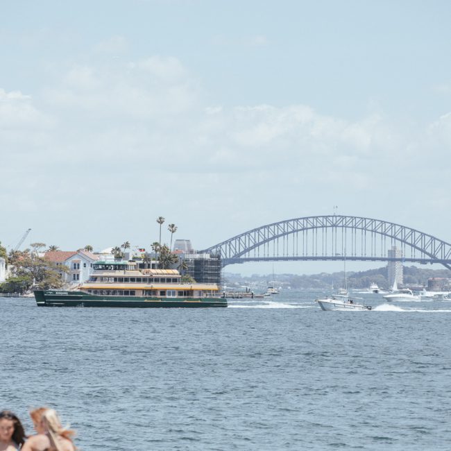 A ferry and several smaller boats navigate the waters near the Sydney Harbour Bridge on a sunny day, with buildings and trees lining the shore in the background. It's an ideal setting for a Sydney boat party hire, offering spectacular views and lively entertainment.