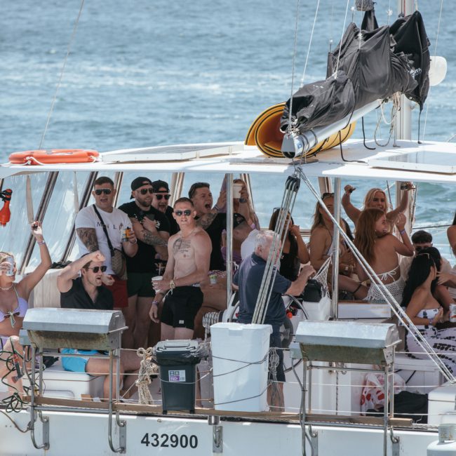 A group of people are gathered on a private yacht charter in Sydney Harbour, socializing and holding drinks. Some are wearing swimsuits and sunglasses. The boat has life vests and a visible identification number "432900".