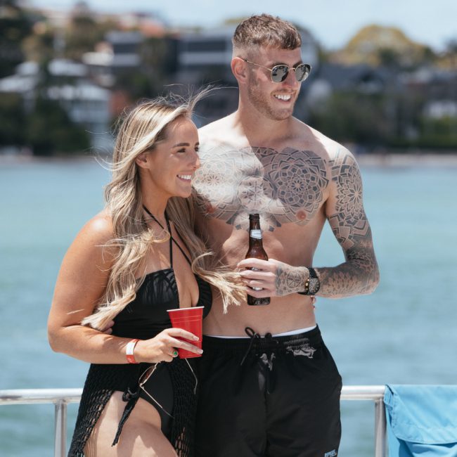 A man and woman in swimwear are standing together on a boat, smiling and holding drinks. A body of water and buildings in the background suggest a coastal setting, perfect for a Sydney boat party hire.