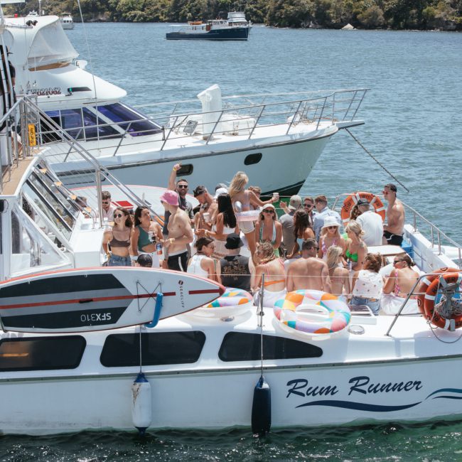 A group of people are gathered on the deck of a white boat named "Rum Runner" on the water, enjoying what looks like a Sydney boat party hire. Another boat is visible in the background. Various floatation rings and a paddleboard are also on the deck, perfect for a private yacht charter in Sydney Harbour.