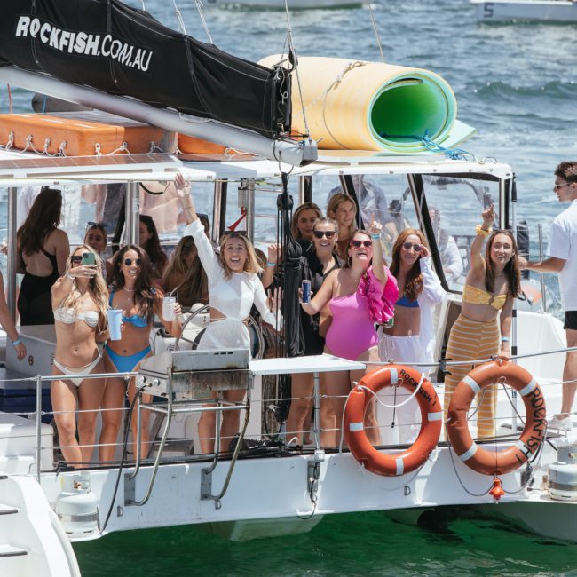 A group of people on a boat enjoy a sunny day while taking photos and socializing. The boat, named 'Rockfish.com.au,' floats gracefully on the water. Perfect for private yacht charters in Sydney Harbour or corporate boat events in Sydney.
