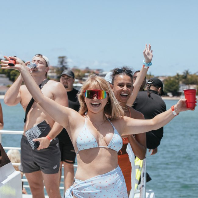A group of people on a boat, smiling and holding drinks. A woman in the foreground wearing a light blue swimsuit and reflective sunglasses has her arms raised in excitement at the fabulous Sydney boat party hire.