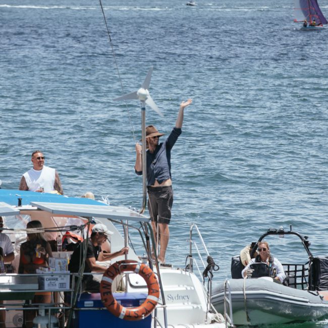A person stands and waves on the back of a boat equipped with a wind turbine, while others sit and watch. Nearby, a small boat with two passengers is seen on the water. Various sailboats are in the background, capturing the essence of Luxury yacht hire Sydney.