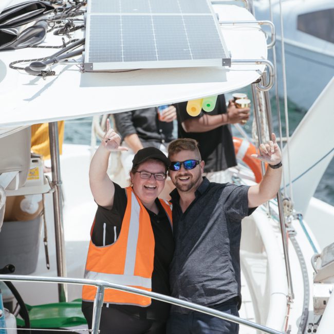 Two people stand on the deck of a boat, smiling as one points upwards. Both wear sunglasses and casual clothes. A solar panel is visible on the boat's roof, suggesting an eco-friendly touch. Others are in the background enjoying what could be a catamaran party in Sydney.