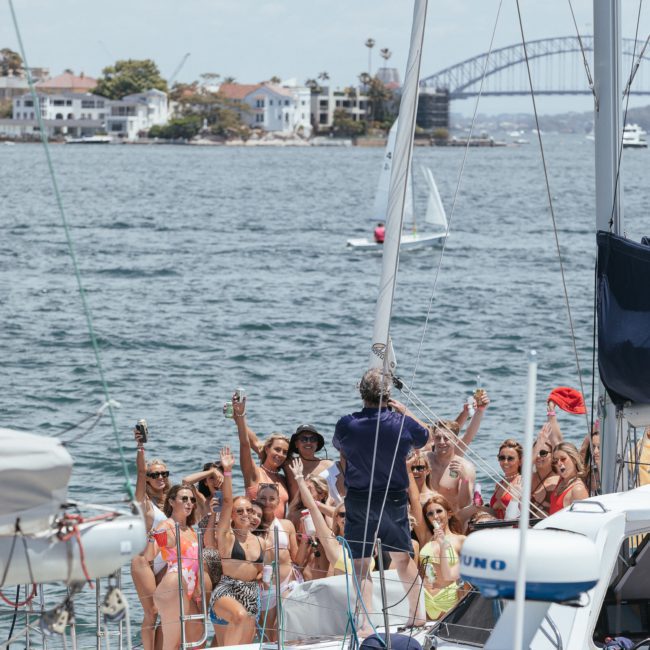 A lively group of people on a boat raise their drinks while a person in the center holds a microphone. Luxury yacht hire Sydney creates an unforgettable experience, with houses and a bridge visible in the background, adding to the picturesque scene.
