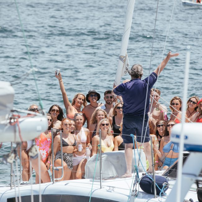 A large group of people in swimwear are gathered on a sailboat in sunny weather, listening to a person at the front. The boat is on water with other boats visible in the background, showcasing the perfect Sydney boat party hire experience.