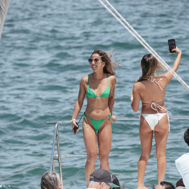 Two women in bikinis are on a boat. One woman in a green bikini is standing and smiling at the camera, while the other in a white bikini is taking a selfie. Others are seated nearby, enjoying their luxury yacht hire Sydney experience.