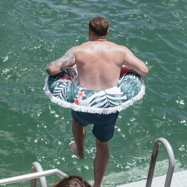 A man with a tattoo on his right arm wears a tropical-patterned inflatable ring, preparing to step into a body of water from a dock. A woman is partially visible in the lower part of the image, hinting at an exciting Sydney boat party hire experience.
