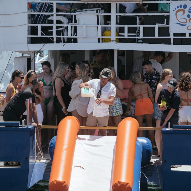 A group of people in swimwear socialize on the deck of a boat with a water slide. Some are standing, and one person is holding a cooler. The luxury yacht "Barefoot Charters" offers an exclusive Sydney boat party hire experience on the stunning Sydney Harbour.