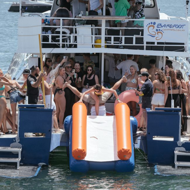 A group of people is enjoying a lively catamaran party on Barefoot Charters, with some using a waterslide at the rear. They are wearing swimsuits and lounging on the deck. The boat is anchored in a body of water, perfect for corporate boat events in Sydney.