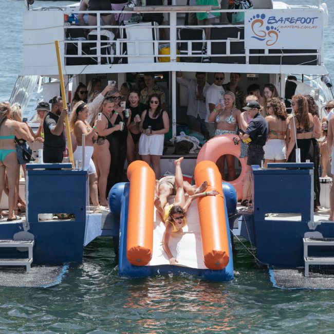 A lively group of people are enjoying a boat party. Two individuals lounge on an inflatable slide leading into the water. The boat, named "Barefoot," is docked on a sunny day, making it the perfect setting for a catamaran party in Sydney or even corporate boat events with DJ boat hire options available.