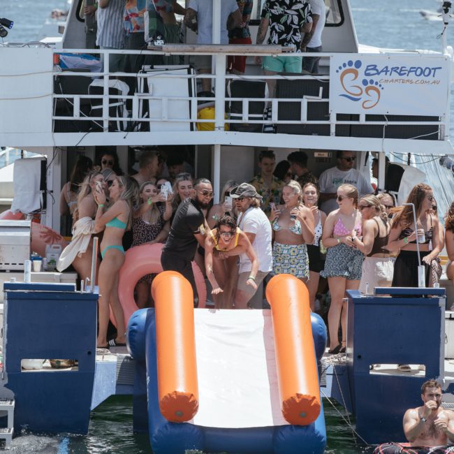 A group of people in swimwear are enjoying a party on a luxury yacht with a slide into the water labeled "Barefoot." Some are on the slide, while others are gathered on the deck, fully embracing the festive spirit.