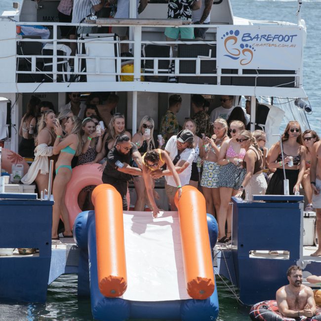 A group of people enjoying a Sydney boat party hire on a luxury yacht with a slide into the water. Various individuals are on the upper deck, and a person is sliding down while others stand and watch.