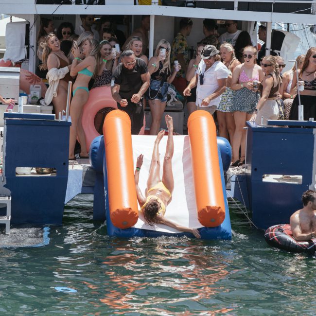 A group of people on a private yacht charter in Sydney Harbour enjoy a party, some watching or socializing, with one person sliding down a dual slide into the water and two people floating on inflatables nearby.