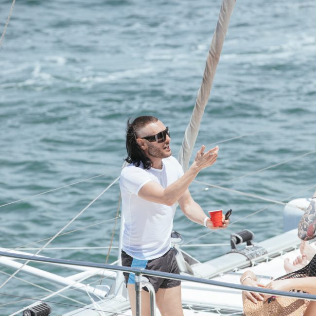 A man with sunglasses and a drink stands on a boat, gesturing with his hand, while another person in a straw hat sits nearby. The ocean serves as the idyllic backdrop for this scene of relaxation on a luxury yacht hire Sydney.