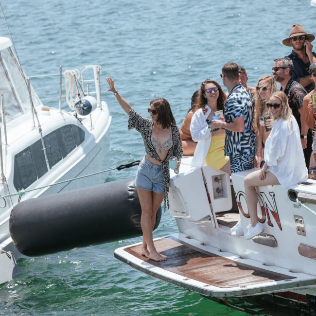 A group of people stands on the deck of a yacht docked alongside another boat in sunny weather. One person is waving while others socialize, some holding drinks, enjoying a private yacht charter on Sydney Harbour.