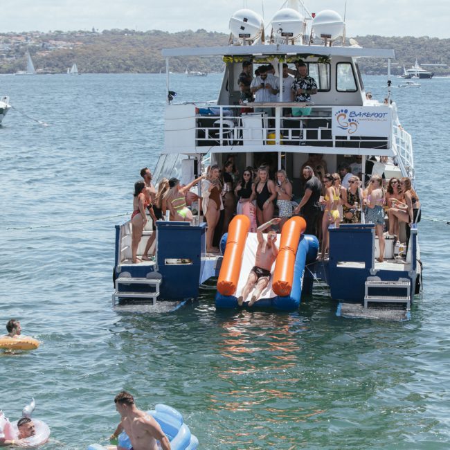 A group of people are on a catamaran party in Sydney with slides, docked in a body of water. Some individuals are using the slides and swimming alongside inflatable tubes and floats, enjoying the vibrant atmosphere.