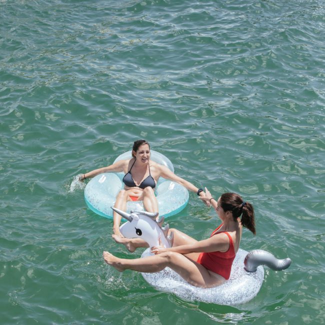 Two women in swimsuits float on inflatable rings in a body of water. One is on a white unicorn float, and the other on a clear ring. Other people are also seen in the water on floats, enjoying what looks like an extension of a lively catamaran party Sydney event.