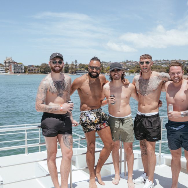 Five men in swimwear stand arm-in-arm on a boat deck, smiling. They are holding drinks, with water and a coastal cityscape in the background. The sky is clear and sunny, perfect for their luxury yacht hire Sydney adventure.