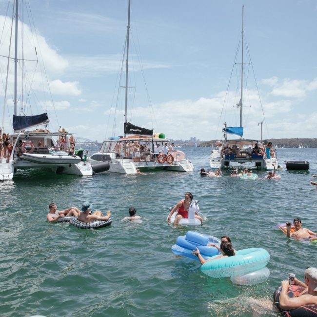 People are enjoying a sunny day in the water near several docked boats. Some are swimming while others float on inflatable tubes and rafts, all amidst a scene perfect for a luxury yacht hire in Sydney Harbour.