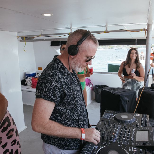 A man wearing headphones is playing music on DJ equipment on a boat, with people standing in the background, enjoying a Sydney boat party hire.