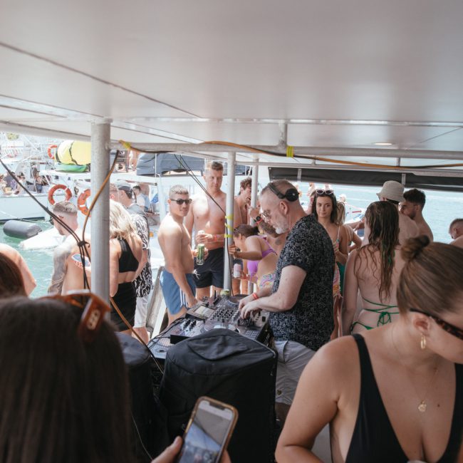 A group of people enjoy a festive atmosphere on a luxury yacht hire in Sydney, with a DJ playing music. Some are dancing, while others are socializing and drinks are visible in the hands of several attendees.