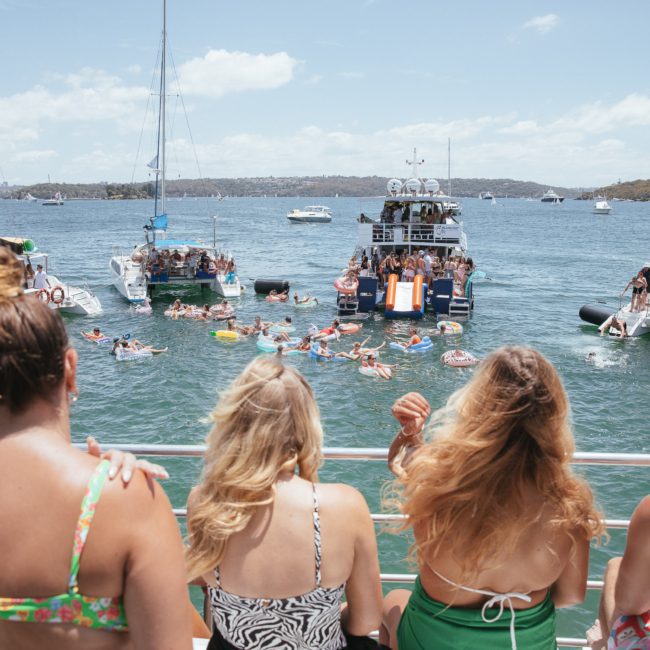 A group of people on a boat enjoy watching others swimming and floating in a sunny bay surrounded by multiple boats and yachts, as part of a Sydney boat party hire.