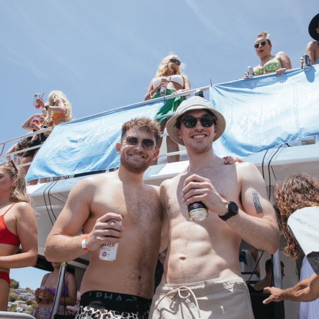 Two men in swimwear stand on a boat holding drinks. Other people in swimsuits are on the upper deck, some looking down at the camera and others interacting among themselves. Experience this scene with a Sydney boat party hire, perfect for a memorable event.