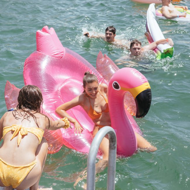 People enjoying a sunny day in the water with inflatable floats, including a large pink flamingo. Two individuals in yellow swimsuits interact on the flamingo. Others swim and float nearby, soaking up the fun of a Sydney boat party hire.